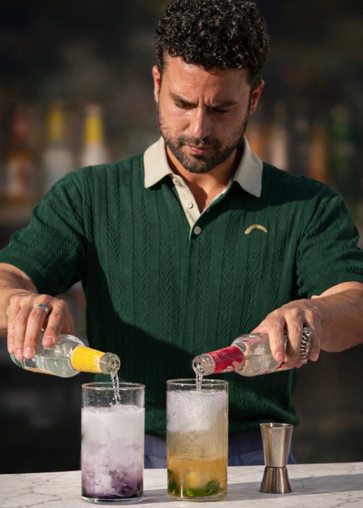 A man in a dark green knit polo sweater pours two clear cocktails into two glasses on a marble counter in a bar setting.
